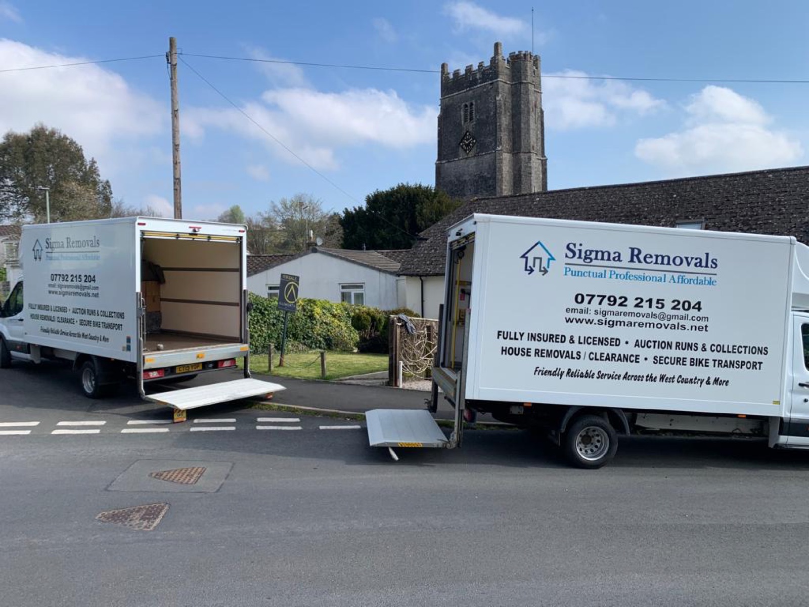 Two branded Sigma Removals Luton vans with tail-lifts down, parked in a Devon village ready to unload